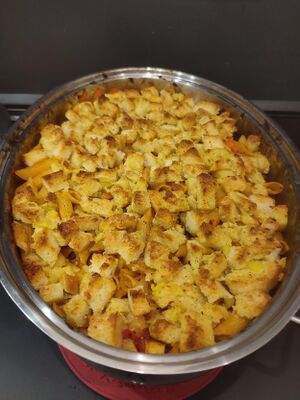 Photo of a casserole inside a large rounded metal casserole dish. The casserole is still intact showing a top crust of browned garlic bread crumbs with some vegetables mostly tomatoes and carrots poking out.